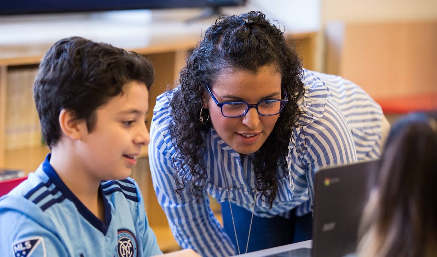 Uma educadora de óculos e um aluno estão observando algo na tela do laptop.