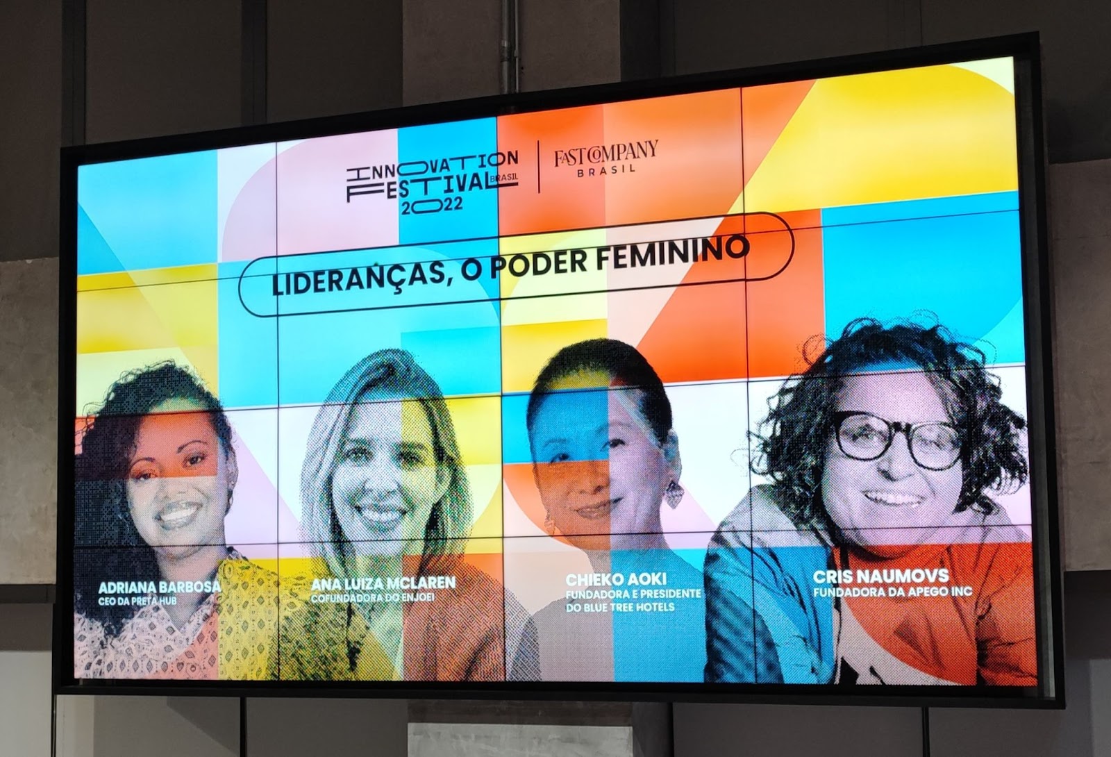 Foto do painel do Innovation Festival 2022 com muitas cores vivas (azul, amarelo, lilás e branco) em formatos geométricos. Abaixo do nome do evento, está  o título “Lideranças, o poder feminino” e a foto das quatro palestrantes, Adriana Barbosa, uma mulher negra de cabelos crespos pretos abaixo do ombro; Ana Luiza McLaren, uma mulher branca de cabelos loiros; Chieko Aoki, uma mulher amarela de cabelos pretos que estão presos para trás; e Cris Naumovs, uma mulher branca de óculos de grau e de cabelos castanhos curtos e cacheados.
