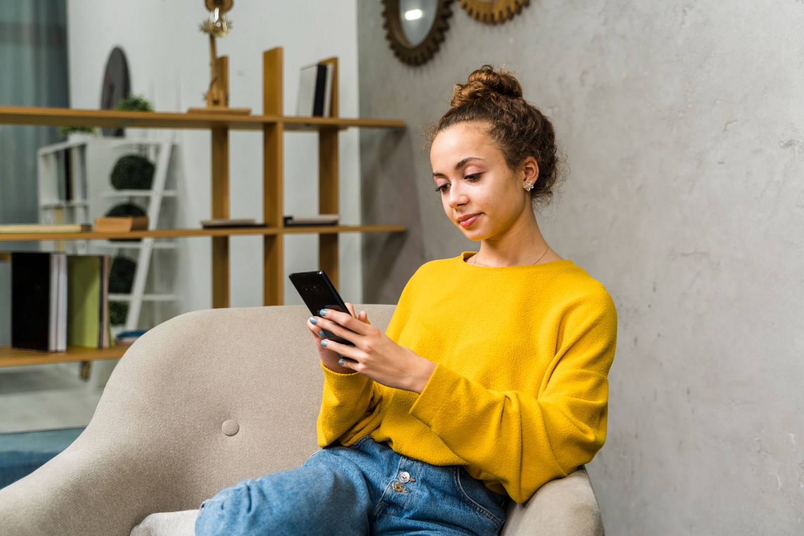 Imagem de uma jovem mulher branca, de cabelos presos, sentada em um sofá enquanto mexe no celular. Ela veste uma blusa de frio amarela e uma calça jeans.