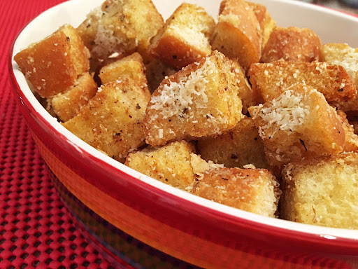 Croutons with cheese in a red serving bowl on a red table mat.
