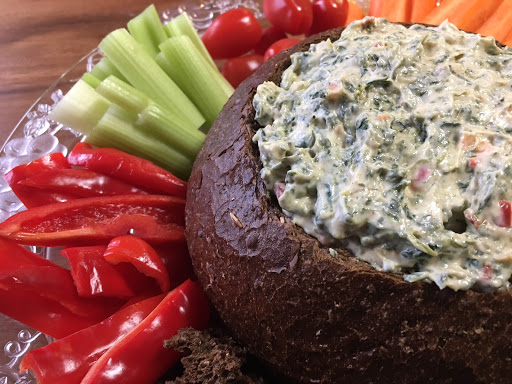 A pumpernickel bread stuffed with a spinach dip on a glass plate with cut veggies around it.