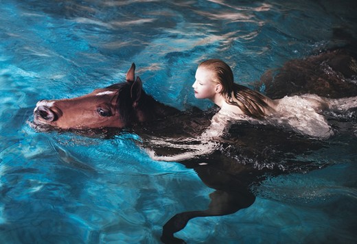 A imagem mostra uma mulher nua nadando. Ela se agarra ao pescoço de um cavalo marrom.