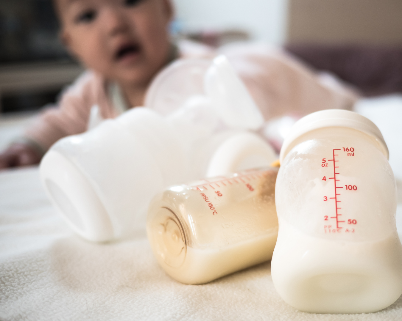 baby lying propped up on the bed staring at breastmilk in a bottle beside a manual breast pump