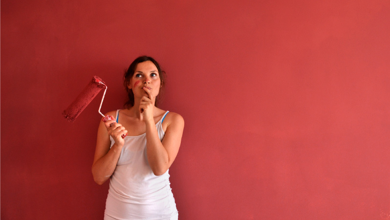 A woman is standing in front of a freshly painted red wall, holding a used paint roller and looking pensive.