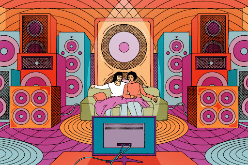 Two women are sitting on a couch are surrounded by speakers and are looking at a TV.