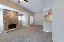 Living room with a brick fireplace, neutral walls & carpet, light trim & a ceiling fan. The entry door is in the background.