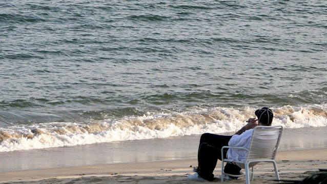 Chorão sentado numa cadeira de praia observando o mar. Está vestido dos pés à cabeça com roupas preto e brancas com as mãos cruzadas em frente ao rosto, parecendo estar refletindo sobre algo.