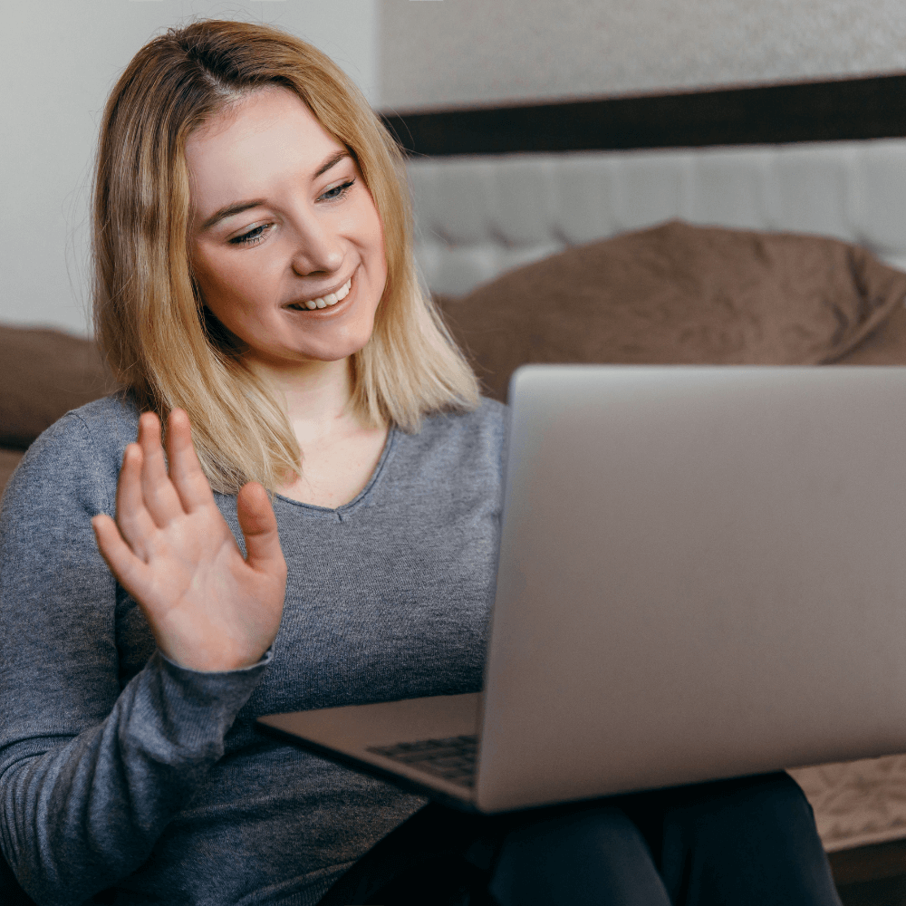 Woman in a teletherapy counseling session smiling and waving at her computer while seated on the floor with her computer resting on her knees. 