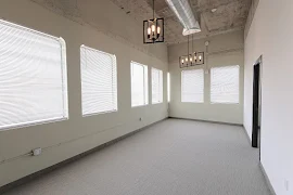 Living area with modern hanging light fixtures, six curved windows with blinds, light carpet and walls, and exposed ductwork