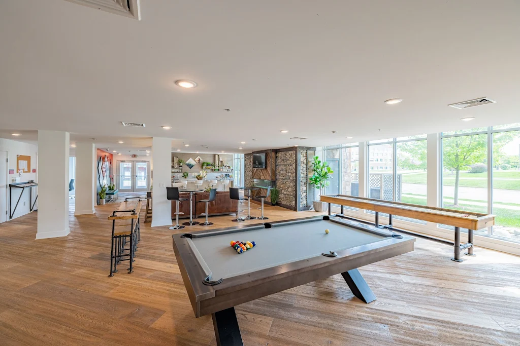 Clubhouse lounge area with pool table, shuffle board, hightop seating, and other lounge areas. Large windows and wood floors.