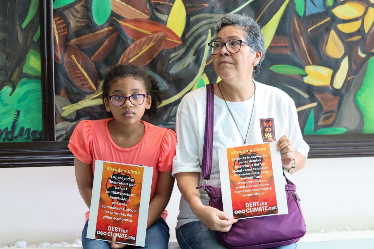 A woman and young girl hold Debt For Climate placards, seated in front of a colourful painting