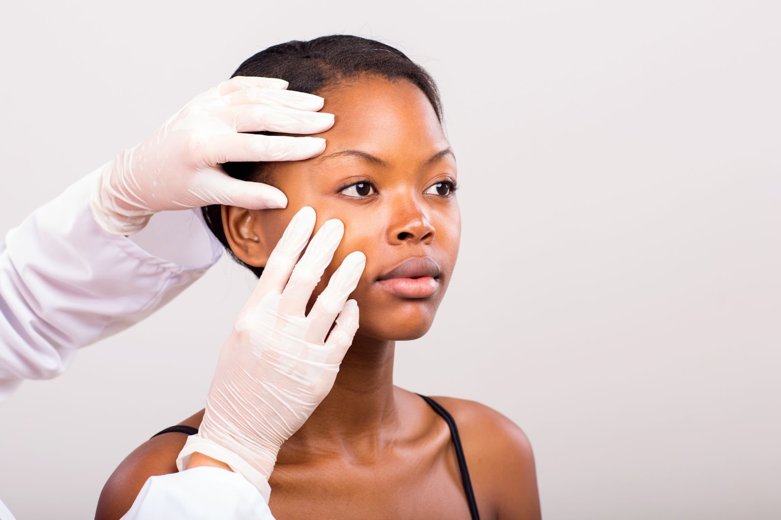 A imagem mostra uma mulher negra em um consultório, tendo sua pele examinada. Ela está de cabelos presos, uma blusa de alças pretas e vemos apenas o recorte das mãos de um médico usando luvas brancas e tocando no rosto dela.