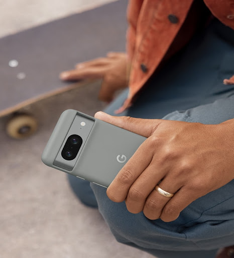 A person kneels beside their skateboard. In their left hand is a Pixel 8 in a Pixel 8 Case. They also wear a Pixel smartwatch.