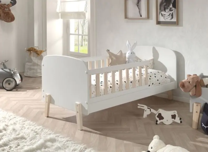 A white toddler bed with rails, surrounded by toys, in a bright room.