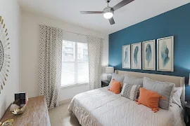 Furnished bedroom with blue accent wall, beige carpet, double window with blinds, and a ceiling fan