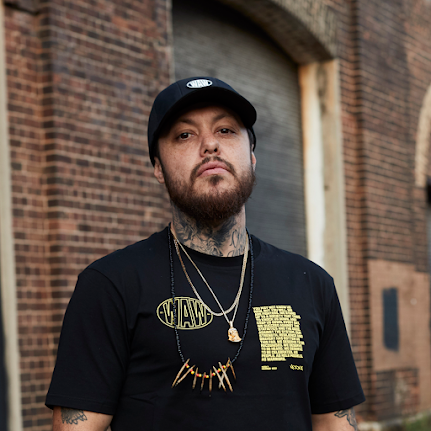 Image of Nooky against a brick wall wearing a black WAW t-shirt with a black cap.