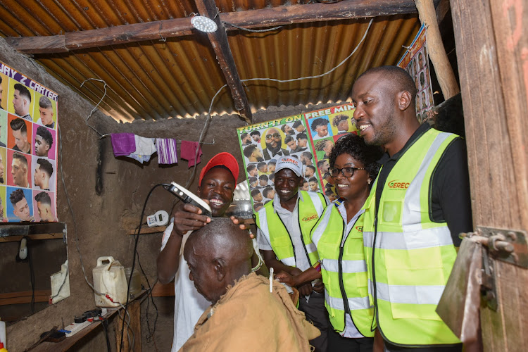 REREC Board Director Milton Lucheri, CEO Dr Rose Mkalama and Director Philip Cherige look on as a customer enjoys a haircut at a barber shop in Kakoldong village, Teso South constituency, one of the projects under the rural electrification programme.