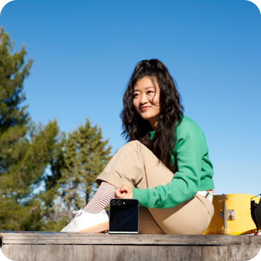 The Samsung Galaxy Z Flip6 is unfolded halfway so that it’s propped upright. It’s facing a person happily sitting outside and carrying out a conversation on a sunny day.