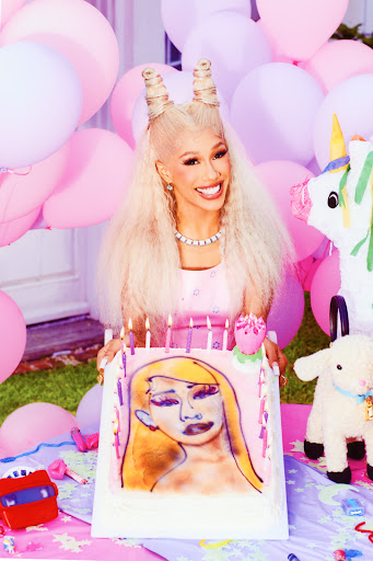 Bia poses with a square birthcay cake airbrushed with her likeness. She is surrounded by balloons and a piñata. 