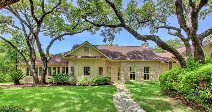 A light-colored residential home in West Lake Hills, Austin, TX. The home has many windows and both brick and siding exterior. Trees cast shade on a lovely green lawn.