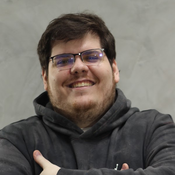 Na imagem vemos o streamer Casimiro sorrindo. Ele é um homem branco com cabelo e barba morena e usa um óculos de grau.