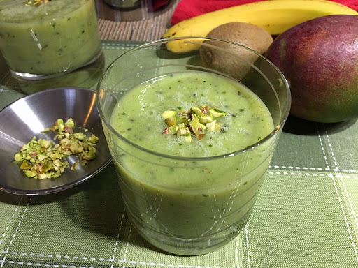 A couple glasses of green smoothie with a small bowl of pistachios and mango, kiwi and banan in the background