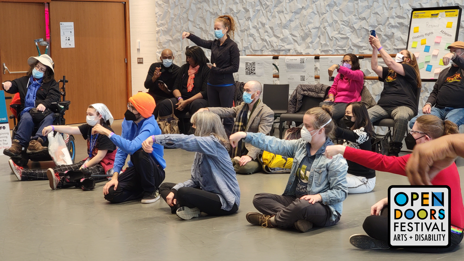 A movement class in a rehearsal room. Twelve participants hold out their arms and pinch their hands downward. They are different ethnicities and wear casual clothing and masks. All are seated, except for one. One participant uses a power wheelchair. The Open Doors Festival logo is in the bottom right corner of the image.