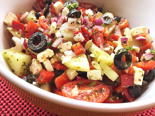 A mixed salad (black olives, tomatoes, cucumber, red peppers, etc.) in a large bowl on a red mat.