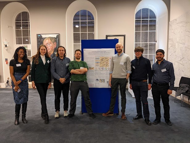 Seven people standing either side of a conference poster board. They are smiling. Three long, round windows are in the background.