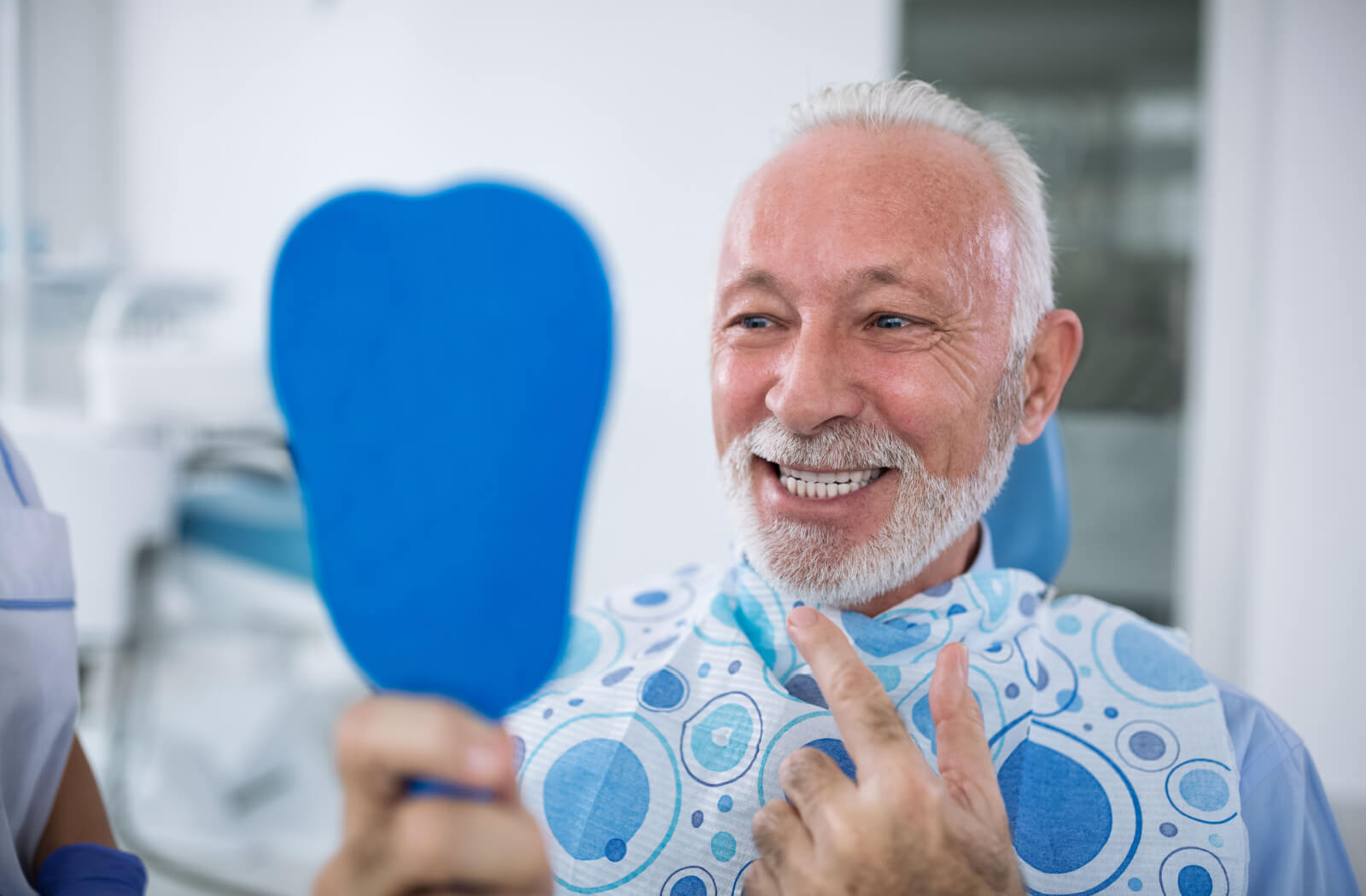 A senior man sitting in a dentist's chair, holding up a mirror and smiling with his dentist