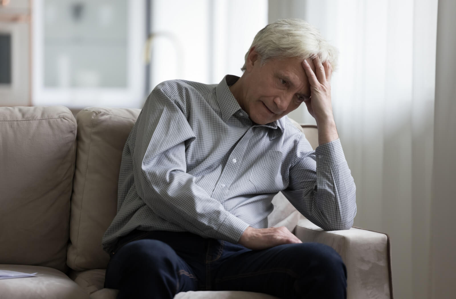 A senior man with a look of confusion and irritability sitting on a couch in a brightly lit room.