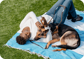 A Black woman lays on a blanket in the grass with her German Shepard service dog while holding her Android in her hand.