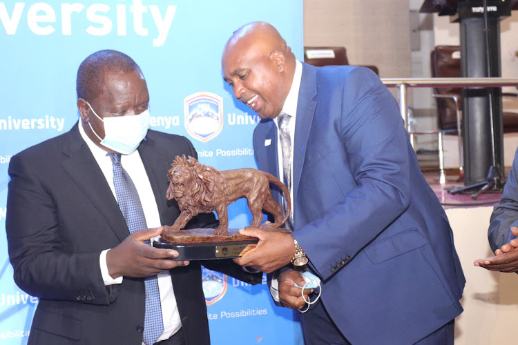 Interior CS Fred Matiang'i receives a gift of a lion curving from MKU Founder Simon Gicharu at MKU main campus in Thika on Thursday.