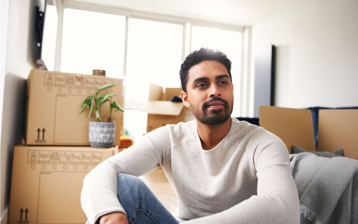 A young man sits on the floor of his New York City apartment, surrounded by moving boxes. 