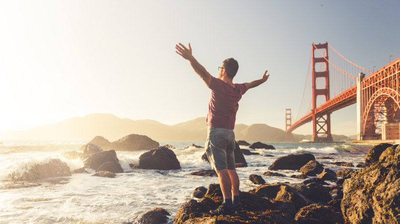 A man is standing on the coast beside the Golden Gate Bridge in San Francisco. His arms are outstretched as he marvels at the view.