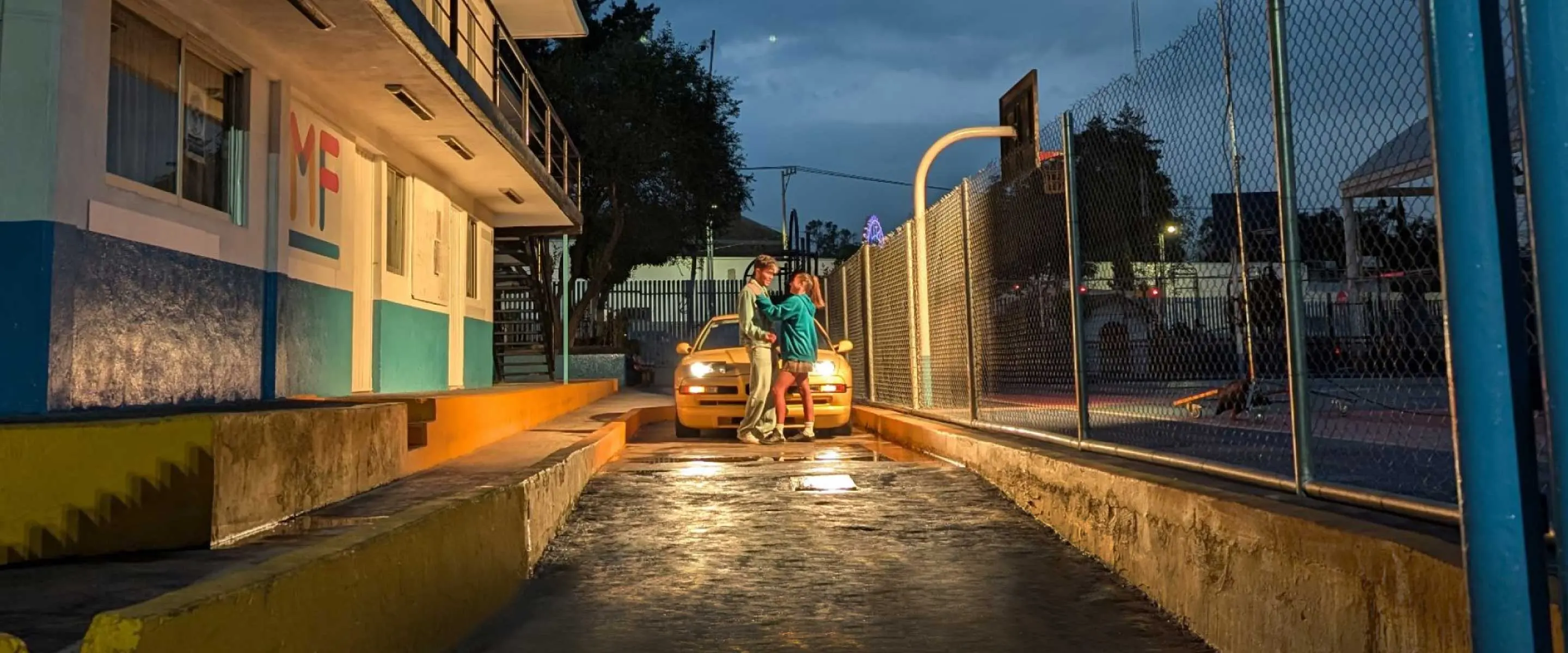 A couple standing in front of a yellow car, outside. 