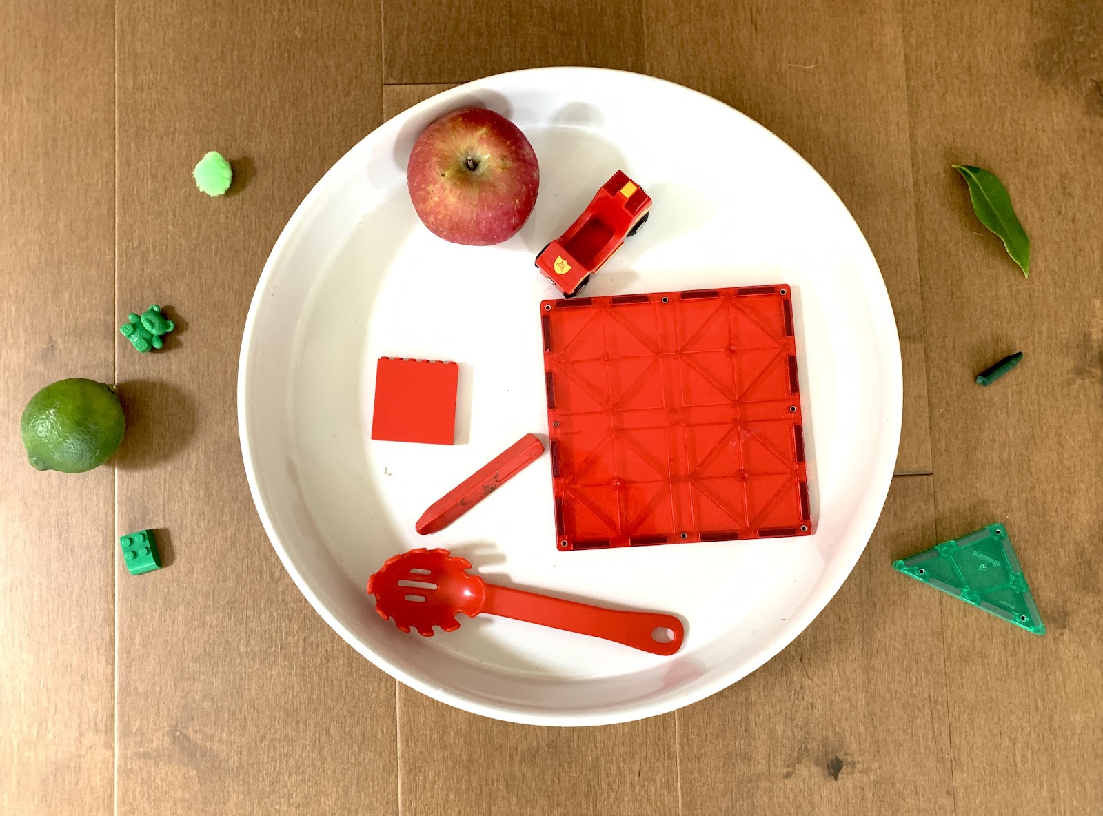 The picture shows a big white circle in the middle. Inside the circle: 1 red apple. 1 big red square. 1 red spoon. 1 red toy car. And 2 more red items. Outside the circle: 1 green building block. 1 lime. 1 green teddy bear. 1 green pom-pom. 1 green triangle. 1 green leaf. And 1 more green item.