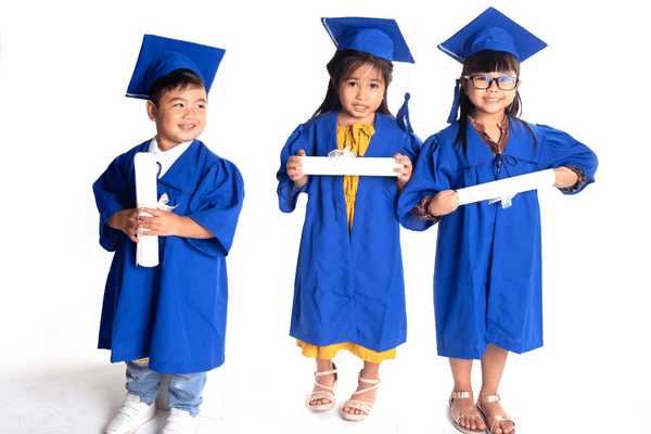 three preschool aged children wearing blue graduation caps and gowns holding rolled, white diplomas