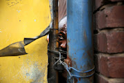 A man suspected of being a foreigner peers through a gap in a fence, while angry locals shout outside.