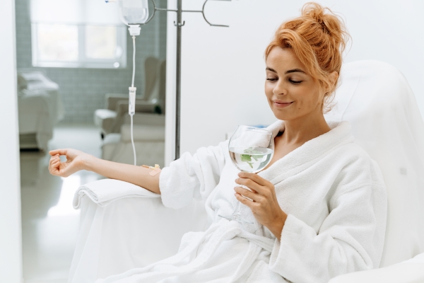 woman-white-bathrobe-sitting-armchair-receiving-iv-infusion-she-is-holding-glass-lemon-beverage-smiling