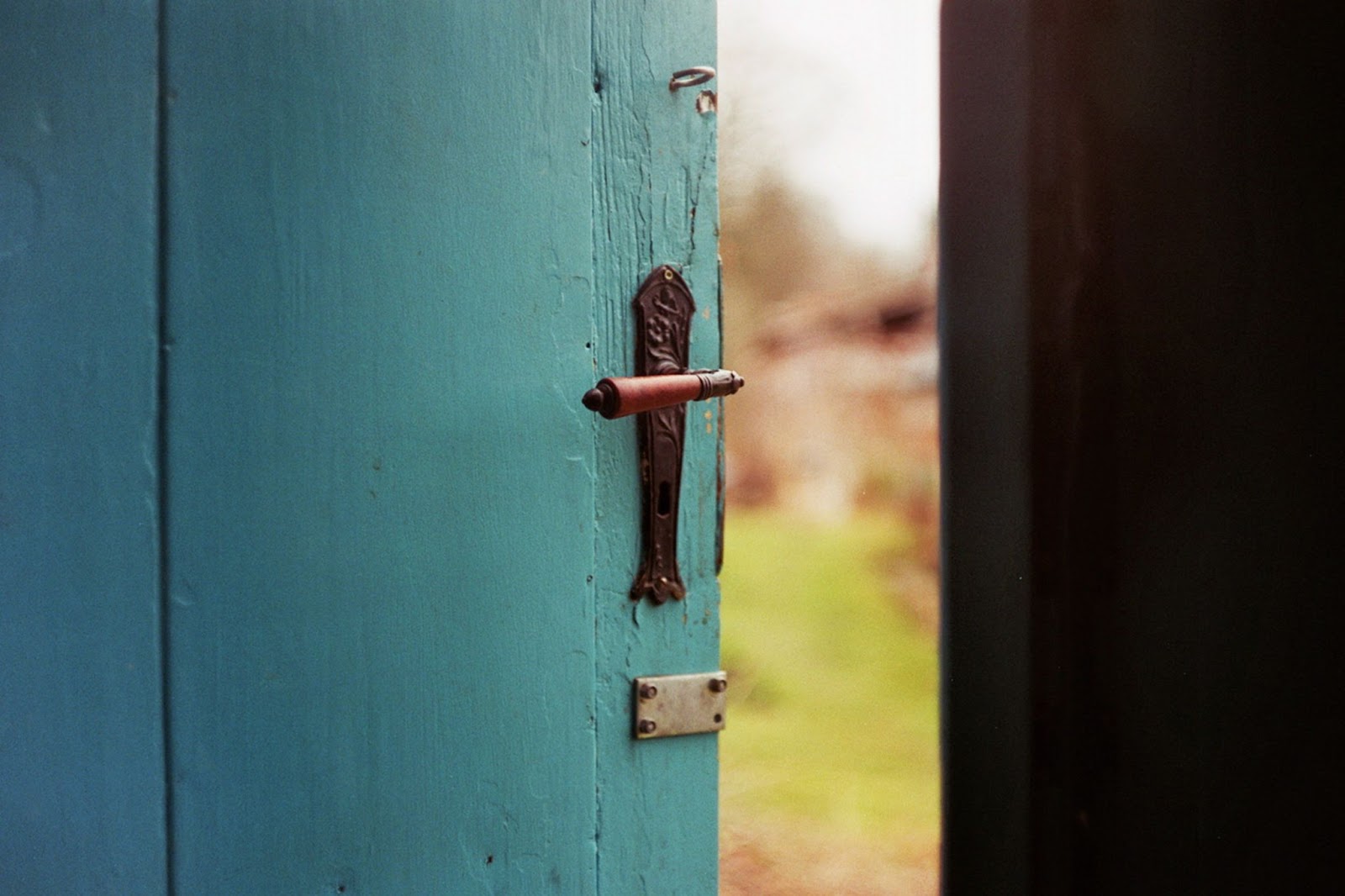 A teal, plank wood door with an ornate, antique handle open just a bit to see an out of focus green path outside.