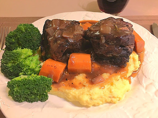 A couple beef short ribs place on top of mashed potatoes along with carrots and steamed broccoli with a nice rich gravy spooned over.