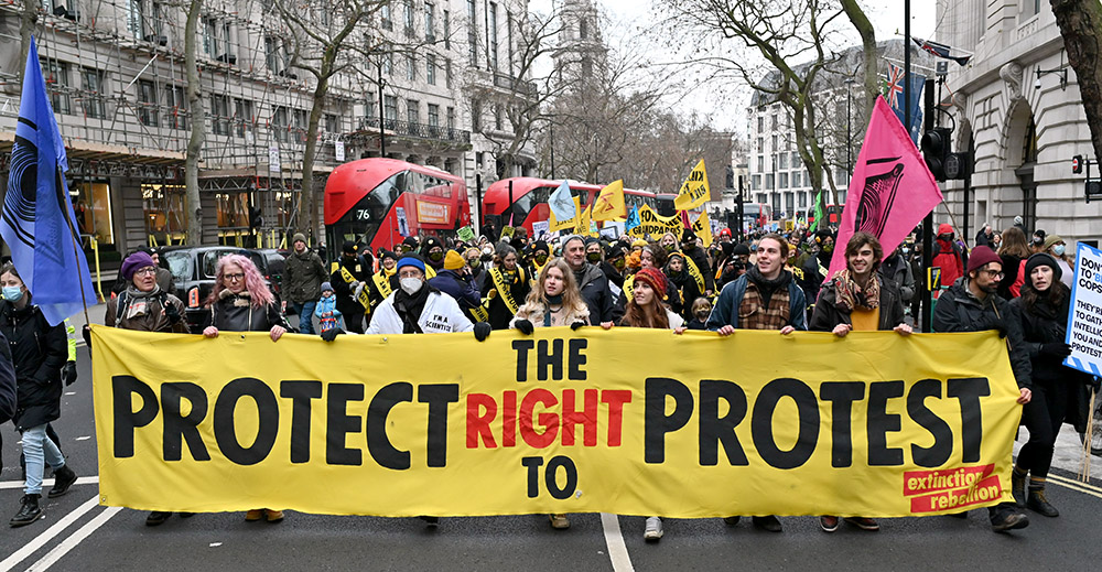 A large column of marchers with a Protect The Right To Protest banner head past London buses on the way to Parliament