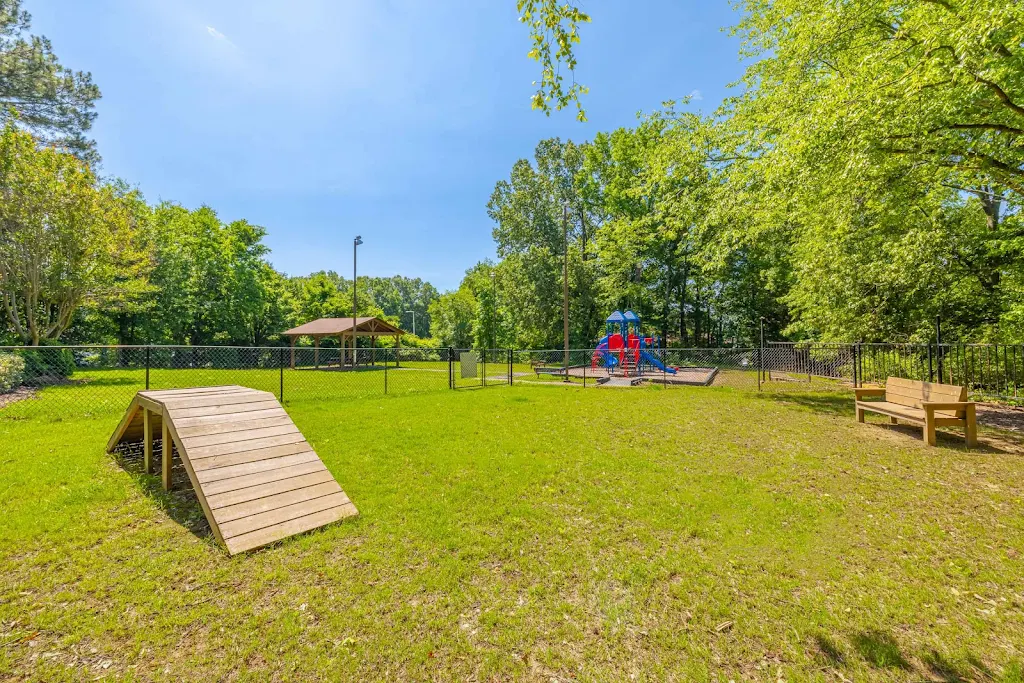 Angled view of fenced-in dog park with wood dog ramp and benches. Playground and covered picnic area in background 