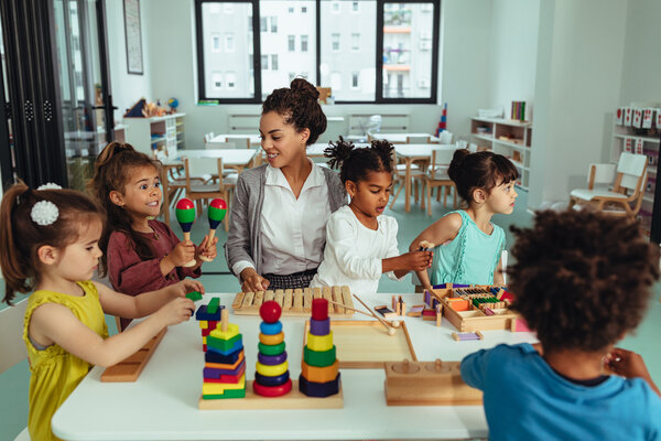 A school teacher is sitting at a table surrounded by five students. The students include four girls and one boy. One little girl with a yellow shirt plays with blocks on the table from right to left. A girl with a red shirt holds Morracos in her hands while the teacher looks and smiles at her. Another girl with a white shirt sits on the teacher's lap and plays, and another girl with a sleeveless blue shirt stares off into the classroom. The little boy's face is not showing, and he is playing with blocks on the table. 