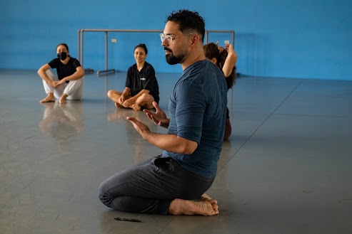 Photo Description: Fernando Ramos is seating with his lower leg tucked underneath his thighs. We see his left profile as he addresses students. He is wearing a long sleeve blue shirt that has been rolled up to the bicep and grey sweatpants. He is a Caribbean man with slightly tan skin, glasses, dark hair and a beard. He has three female students next to him making part of a circle. They are seating on grey linoleum floor. The wall behind them is a bright blue with some visible grey ballet barres stacked against it.