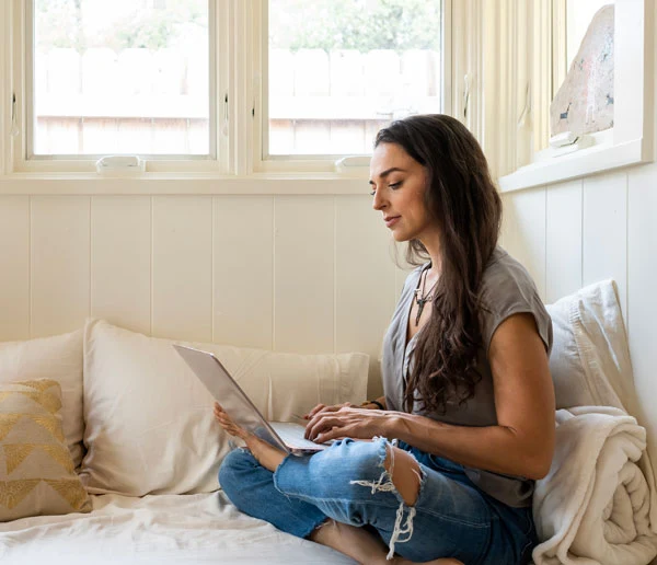 A woman is sitting on her bed with her laptop and browsing the internet