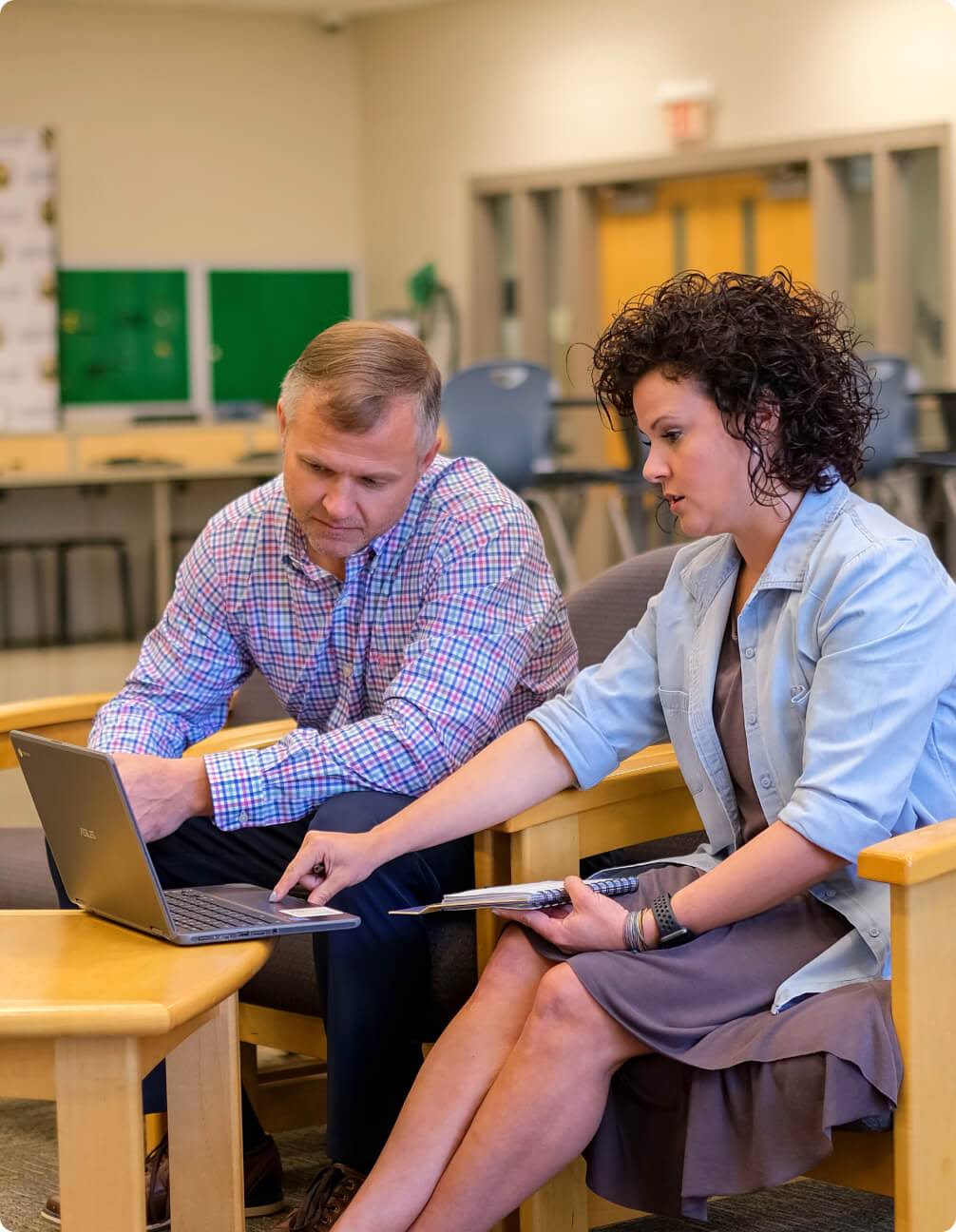 Two people look at a laptop while one touches the trackpad and holds a notebook.