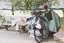 man leest een boek op openbare bank, een been over een zak met bezittingen en geleund tegen een overdekte, volle, bakfiets