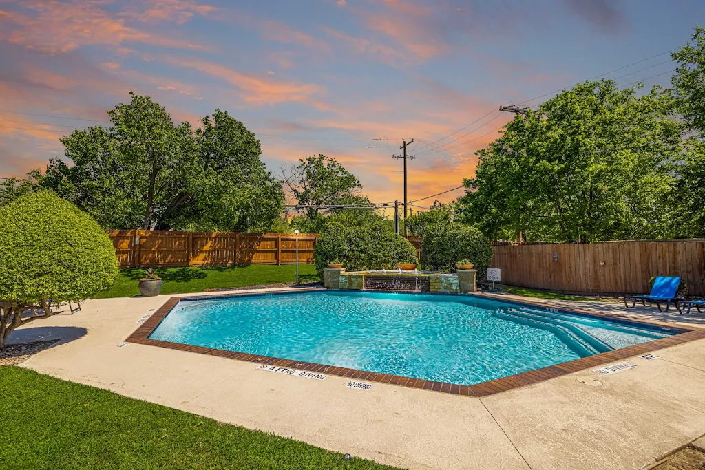 Apartment complex communal outdoor pool with lounging chairs, surrounded by a wooden fence and greenery under a sunset sky.